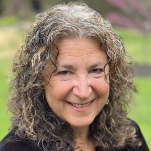 photo of white woman smiling with grey curly hair and black shirt with green meadow behind her